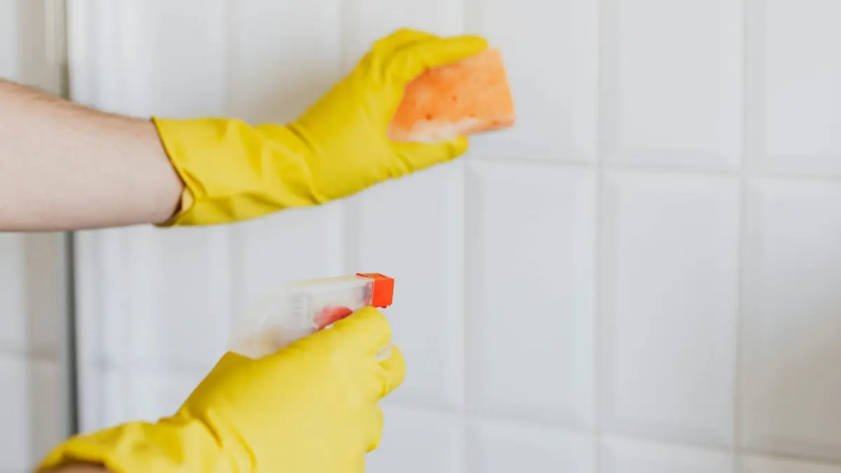 Person wearing yellow rubber gloves cleaning bathroom tiles with a sponge and spray bottle — DIY mold removal is a safe alternative to professional mold remediation cost for surface mold under 10 square feet on non-porous materials